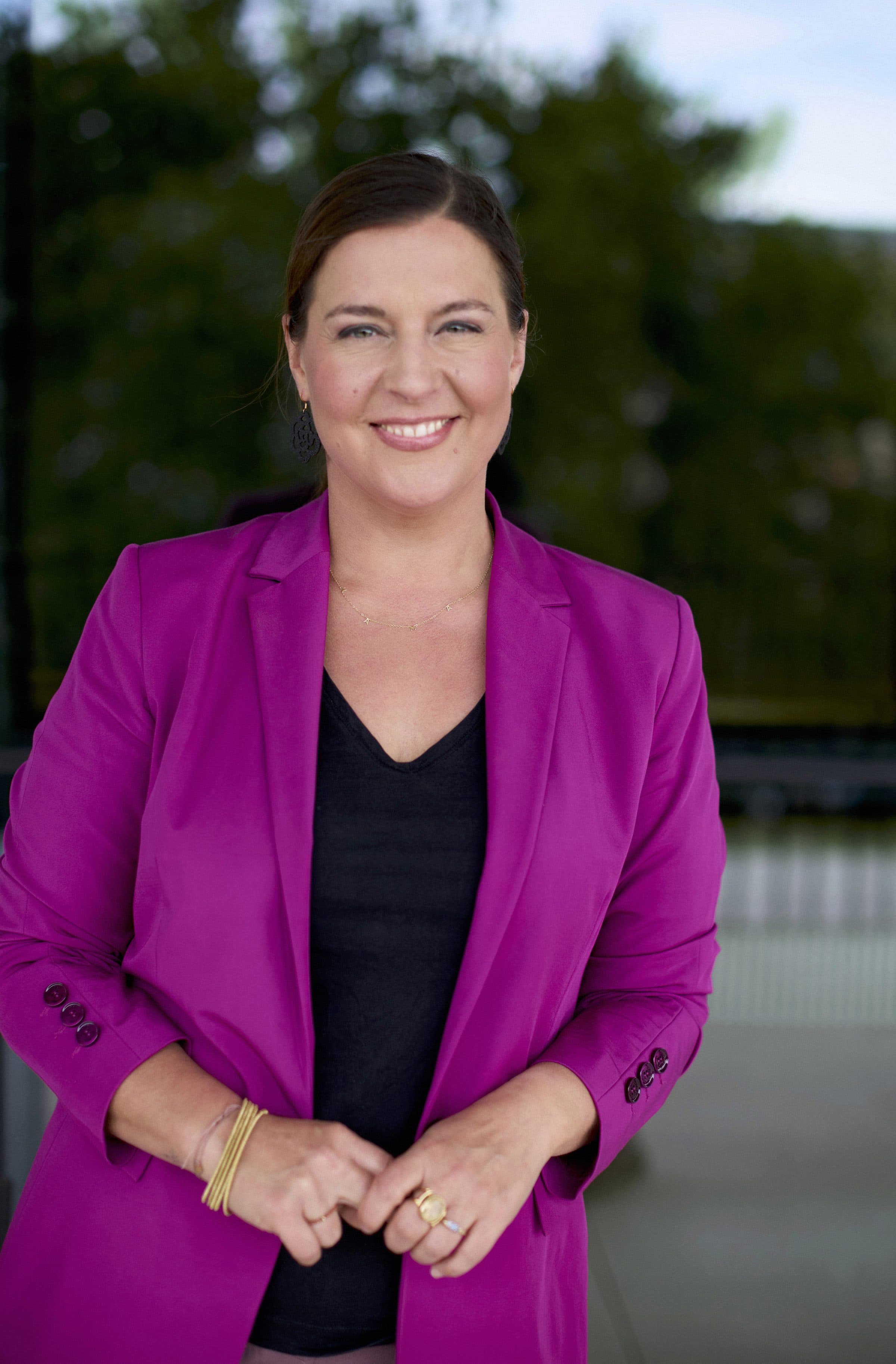 Nele Traen im Magenta-Blazer vor einer Glasfassade, herzlich lächelnd
