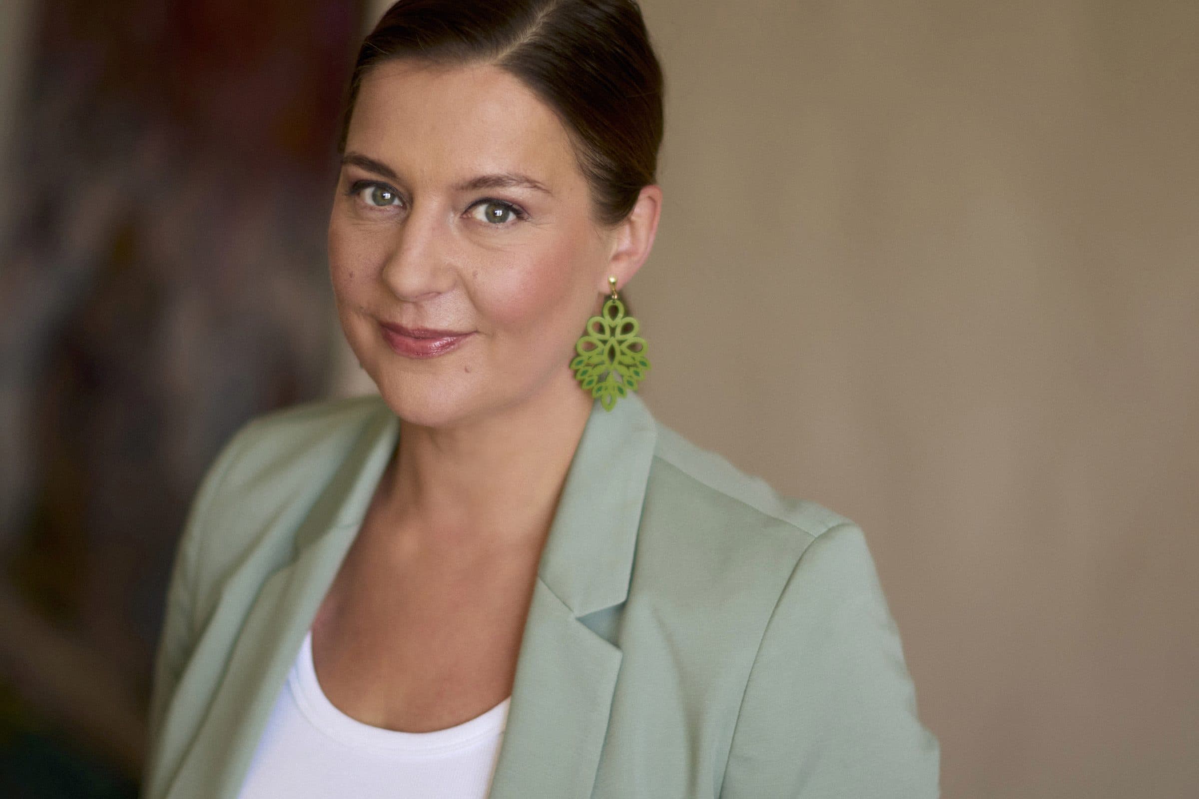 Porträt von Nele Traen im salbeigrünen Blazer mit grünen Ohrringen, warmer direkter Blick
